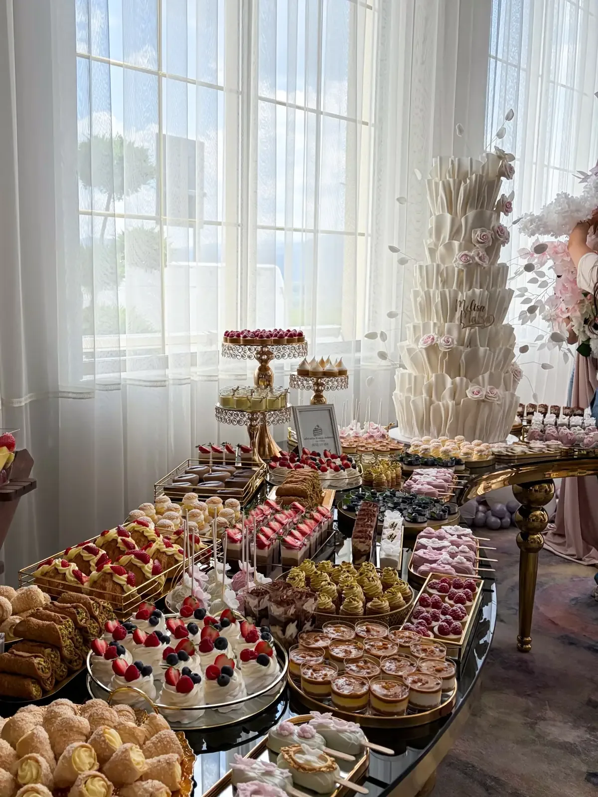 Masă de candy bar cu tort etajat alb cu trandafiri roz, choux, tarte cu fructe și verrine, pe platouri aurii